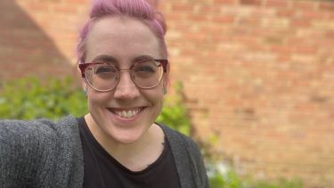 A woman with short pink hair smiles at the camera. She wears a grey cardigan and black top. There is a brick wall and hedges in the background.