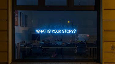Neon sign in a shop window with the text "WHAT IS YOUR STORY?"