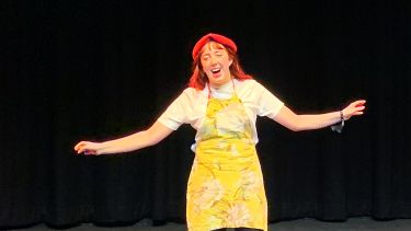 Student with red baret and pinafore arms stretched to the side and mouth open, acting in a play