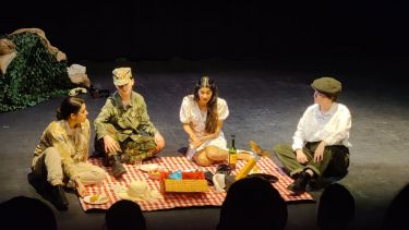 four students on a picnic blanket on a stage. 3 dressed as soldiers