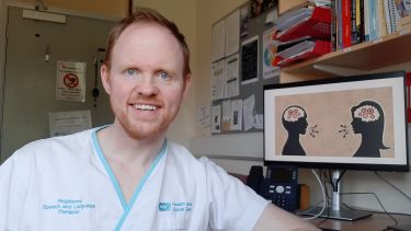 Paddy Moriarty wears white scrubs and smiles at the camera. He has red hair and a short beard. In the background are books, a noticeboard and a computer screen with an image of two cartoon people talking.