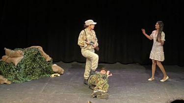Three actors on stage, two dressed up as soldiers, one with a foot on the stomach of the other who is lying on the floor. Woman walks in from the right with arms out. 
