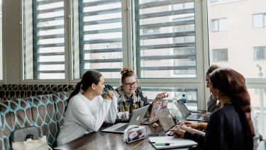 Photograph of four girls sat at a booth table by a window, there are laptops on the desk and they're engaged in conversation