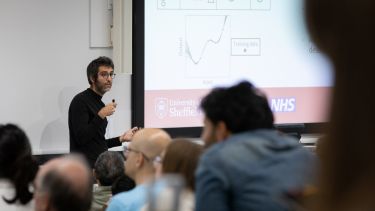 Male speaker with dark hair and glasses wearing a dark polo neck top presents research to an audeince in a lecture theatre