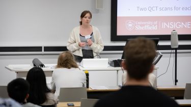 Female speaker  with brown hair tied in a ponytail wearing a light cardigan and striped top speaking to the audience in a lecture theatre