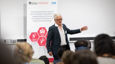 Male professor with short white hair wearing glasses and dark suit with white shirt stands to speak in front of an audience in a lecture theatre in front of a bannerstand displaying red hexagons