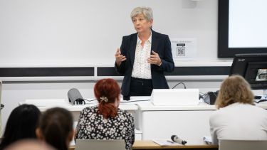 Female professor with short grey hair, wearing a dark suit speaking to the audience in a lecture theatre.