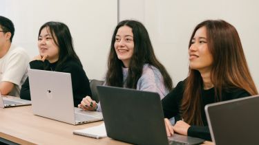Three postgraduate students sitting with laptops, smiling and engaged in a seminar.