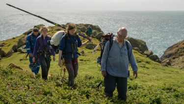 Professor Tim Birkhead walking on a cliff edge on Skomer Island with a film crew behind him