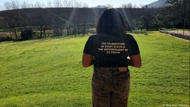Person standing with their back to the camera in a shirt that reads 'the foundation of every state is the empowerment of its youth'