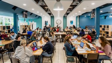 A large room with lots of long rectangular tables with students sat at them with laptops, there is a large clock on the wall 