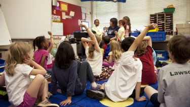 A classroom based lesson with the teacher in the foreground and children holding their hands up