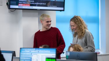 A boy with bleached hair and a red jumper and a woman with curly blonde hair laughing. they are in a newsroom surrounded by computers and TV screens.