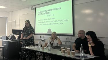 Four people at a panel event. Gemma horton presents at a lectern as three other speakers sit at a long table beside her. 