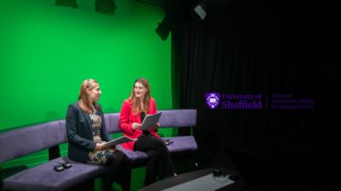 Two girls with blonde hair, holding papers, sitting in a TV studio. School of Journalism, Media and Communication.