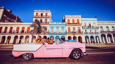 A pink convertible carrying 4 girls drives in front of a strip of brightly coloured houses