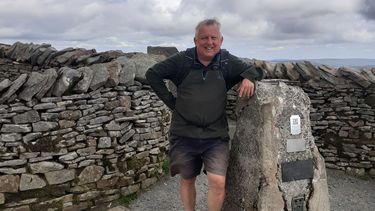 Peter in his hiking outfit stood next to a trig point