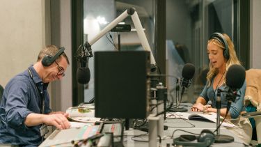 A man and a woman sitting in a radio studio, both wearing headphones. The woman is reading from a script and speaking into the microphone. 