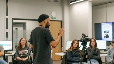 A man in a black beanie speaking to a circle of people, with a camera on a tripod in front of him. 