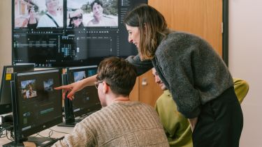 A woman points at a computer screen showing a video editor, as two boys look on.