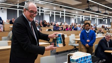 An image of Will Hutton cutting the ceremonial cake baked by Dr Rahul Mandal, who stands behind.