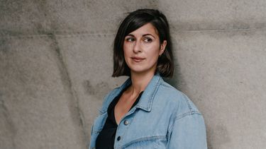 writer Tülin Erkan leaning against a wall, dressed in a jeans jacket and shoulder length dark hair