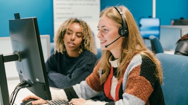 two students at a PC one has a headset on a call the other is talking them through the steps whilst looking at the PC.