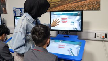 A female university student wearing a headscarf shows a sixth form student in uniform how to use a VR screen.