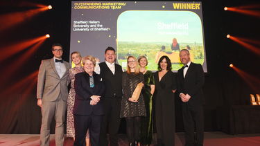Photograph of eight people stood on stage smiling - one of the people holds the THE Award for outstanding marketing and communications team
