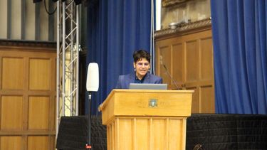Surabhi behind a lectern welcoming the audience to the lecture. There is a covered piano and blue curtains be hind Surabhi. 
