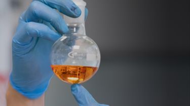 A scientist wearing blue gloves holding up a test tube with an orange liquid inside.