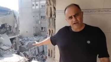 A man wearing a black shirt stands pointing at the rubble of a destroyed city.