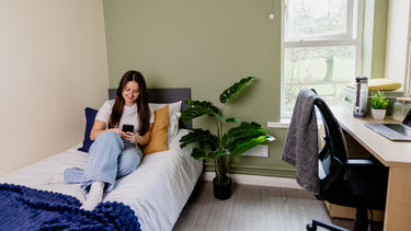 A brown haired girl wearing a white t-shirt and blue jeans sat on a bed looking at her phone