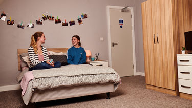 Two girls sat on a double bed, there is a string of photographs hanging from the wall behind them 