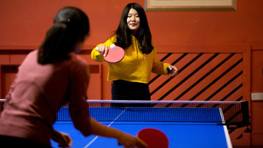 A woman in a yellow jumper holding a paddle in the process of hitting a return shot in a table tennis match
