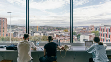 Three students sat at a desk with their backs to the camera. They're facing a view of the Sheffield metropolis from high up.