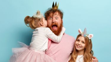 A father a ginger beard, wearing a plastic crown, hugs two small girls. One is in a tutu and the other is dressed as a unicorn.