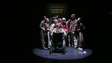 group of actors in a spotlight looking in. In the background it reads Bezoek is een intermezzo