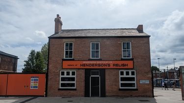 Red brick two storey house with chimney 3 upstairs windows and 2 downstairs windows either side of a black door.  A large rectangular orange Henderson's relish sign sits between the windows of the ground and first floors.