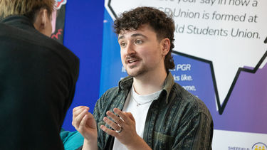 A student with curly hair and a mustache sitting at an activity fair stand. 