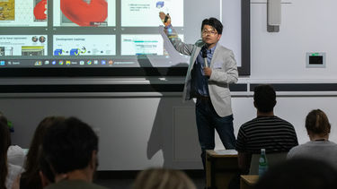 A man with black hair and glasses wearing a blue shirt and light coloured jacket is stood in front of a presentation gesturing and holding a microphone.