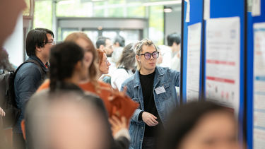 Am man with long bleached blonde hair and dark rimmed glasses, wearing a denim jacket and black top is stood in a crow of people looking at a poster displayl
