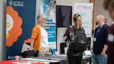 I woman with short grey and dark rimmed glasses and wearing an orange cardican and white top is stood at an exhibitor table with bannerstands behind her talking to a man and a woman