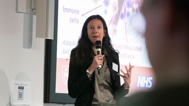 A woman with long dark hair wearing a dark jacket and a brown/beige silk shirt is stood in front of a presentation speaking. She's holding a microphone and gesturing with her other hand.