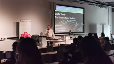 A man with dark hair wearing a white shirt is stood in front of a lectern speaking to a lecture theatre audience and holding a microphone in fron of a presentation about digital twins