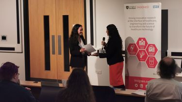 A woman with long dark curly hair wearing a black jacket and white top is smiling and holding a certificate facing a woman with long dark curly hair wearing a dark jacket and red skirt holding a microphone at the front of a lecture theatre in front of an audience
