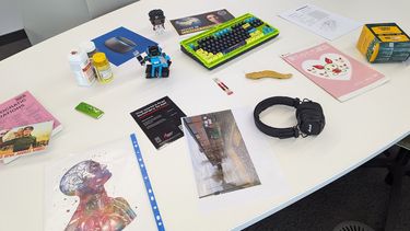 A table with various papers on it as well as a pair of headphones, a small robot, a computer keyboard and some pill bottles