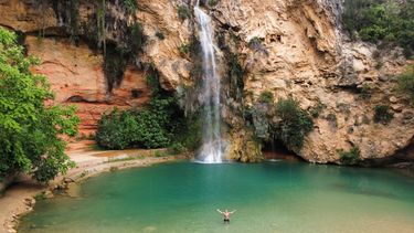 waterfall in spain with a green looking lake in front of it with a student in the water with arms spread out wide