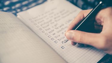 A hand holding a pen writing in a notebook. The notebook has a list of sentences with boxes next to them - a checklist.