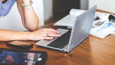 A woman sat at a desk scrolling on the trackpad of a laptop. There is a pile of books, an Ipad and a smartphone on the desk.